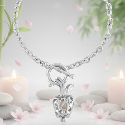 Elegant silver apple-shaped pendant necklace with a pearl inside displayed in a serene spa-inspired setting with candles, stones, and pink petals.