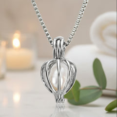 Elegant silver cage-style pendant necklace featuring a central pearl displayed with soft candlelight, a rolled towel, and greenery.