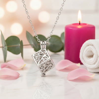 Elegant silver filigree cube pendant necklace displayed with soft pink candlelight, rose petals, and eucalyptus accents.