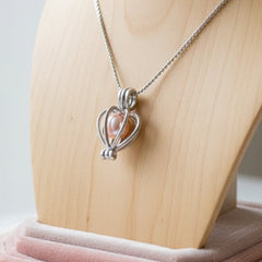 Elegant silver heart-shaped cage pendant necklace featuring a pink pearl displayed on a wooden jewelry stand.