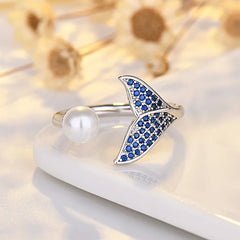 A silver-tone ring shaped like a mermaid tail, decorated with small blue stones, and accompanied by a round white pearl on the band. The ring is displayed on a white ceramic dish with blurred dried flowers in the background.