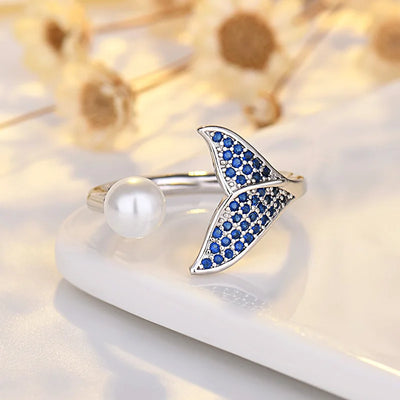 A silver-tone ring shaped like a mermaid tail, decorated with small blue stones, and accompanied by a round white pearl on the band. The ring is displayed on a white ceramic dish with blurred dried flowers in the background.