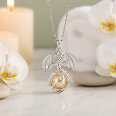 A silver dragon pendant necklace with a pearl inside, displayed on a marble surface with white orchids and lit candles in the background.