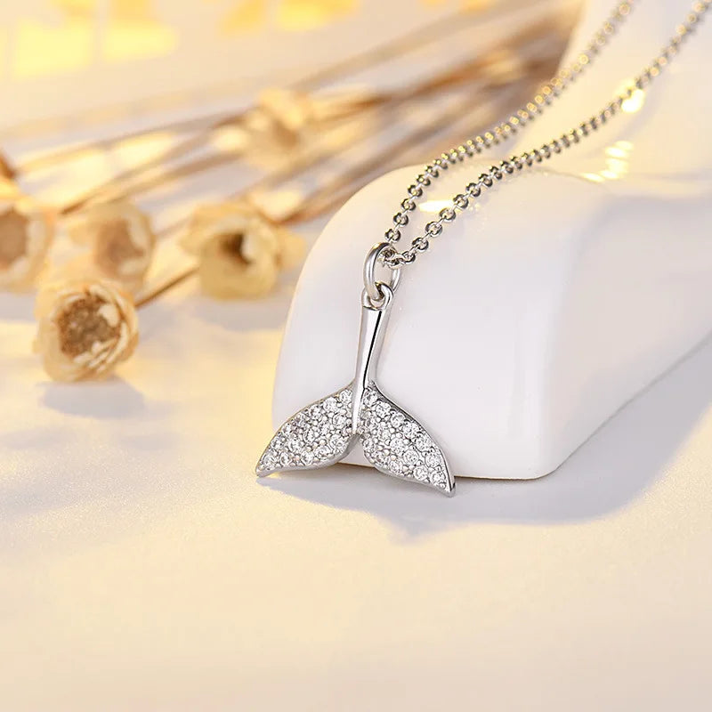 A silver mermaid tail pendant necklace covered in small clear crystals, resting on a white display stand with dried flowers in the background.