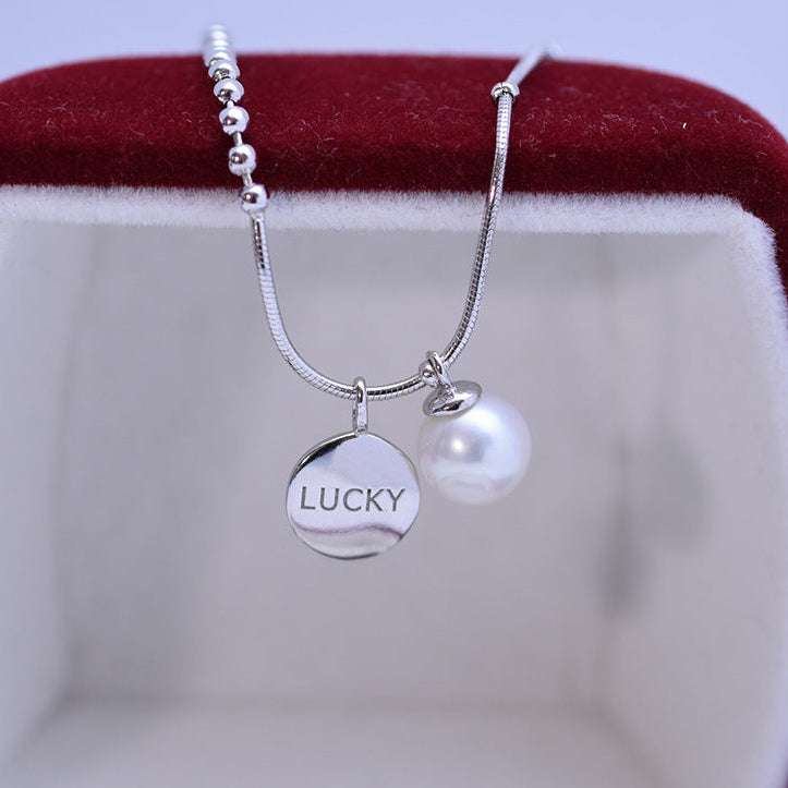 A silver snake chain necklace with a round engraved 'LUCKY' disc charm and a white pearl bead, displayed inside a red velvet jewelry box.