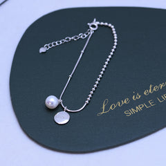 A silver chain bracelet with a single white pearl and a round silver disc charm engraved with 'LUCKY', displayed on a dark green card with gold text.
