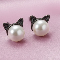 Pair of stud earrings featuring natural freshwater pearls with small black cat ears displayed on a soft pink background.