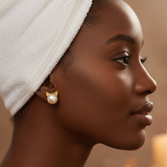 Side profile of a person wearing 925 sterling silver cat‑ear earrings featuring a natural freshwater pearl as the face and small golden ears, styled with a towel wrap and soft cream background.