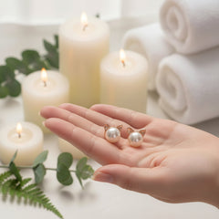 Hand holding a pair of freshwater pearl cat‑ear stud earrings with metallic ears, shown in a serene spa‑style setting with candles, towels, and green leaves in the background.
