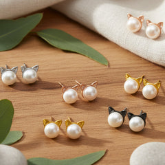 Six pairs of freshwater pearl cat‑ear stud earrings in silver, rose gold, yellow gold, and black styles arranged on a wooden surface with white stones, green leaves, and soft fabric accents.