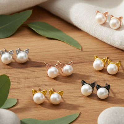 Six pairs of freshwater pearl cat‑ear stud earrings in silver, rose gold, yellow gold, and black styles arranged on a wooden surface with white stones, green leaves, and soft fabric accents.