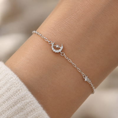 A delicate silver chain bracelet with a crescent moon and star charm, worn on a person's wrist over a cream-colored knit sleeve.