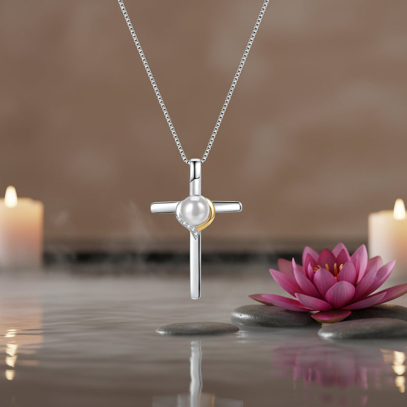 A sterling silver cross pendant necklace featuring a white pearl and a small gold crescent accent, suspended from a delicate silver chain against a spa background.