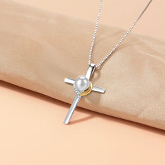 A silver cross pendant necklace with a white pearl and a small gold accent, displayed on a beige satin fabric against a tan background.