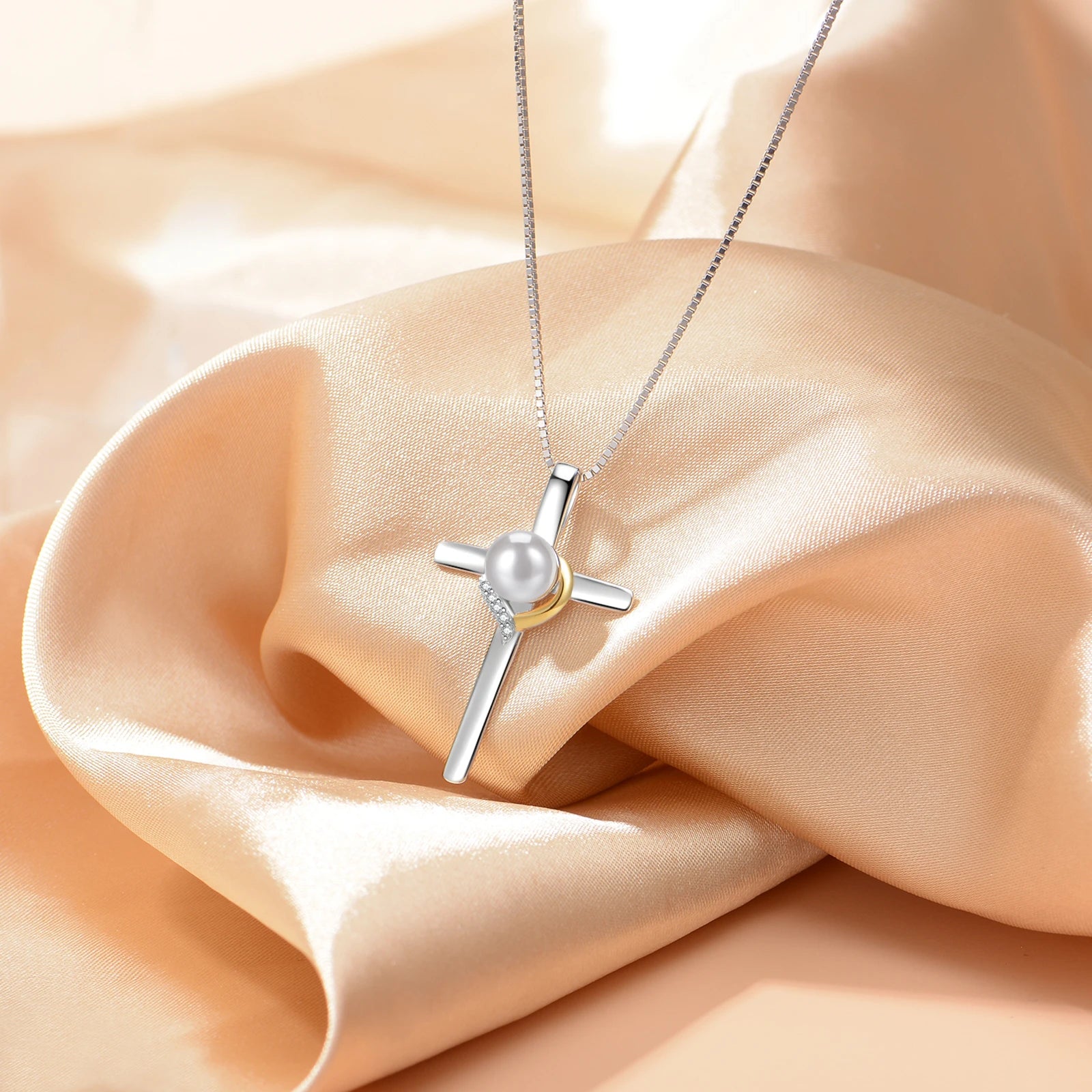 A silver cross pendant necklace with a white pearl and a small gold accent, displayed on draped champagne-colored satin fabric.