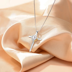 A silver cross pendant necklace with a white pearl and a small gold accent, displayed on draped champagne-colored satin fabric.