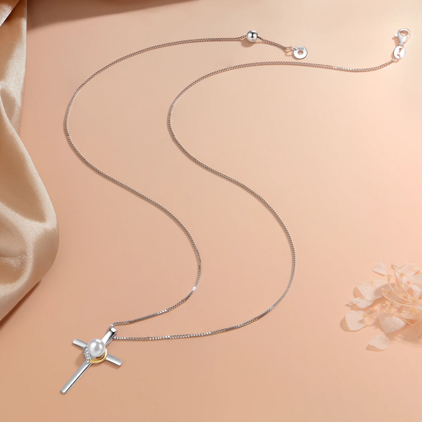 A silver cross pendant necklace with a central white pearl, displayed on a peach-colored background with a satin fabric prop and a decorative flower.