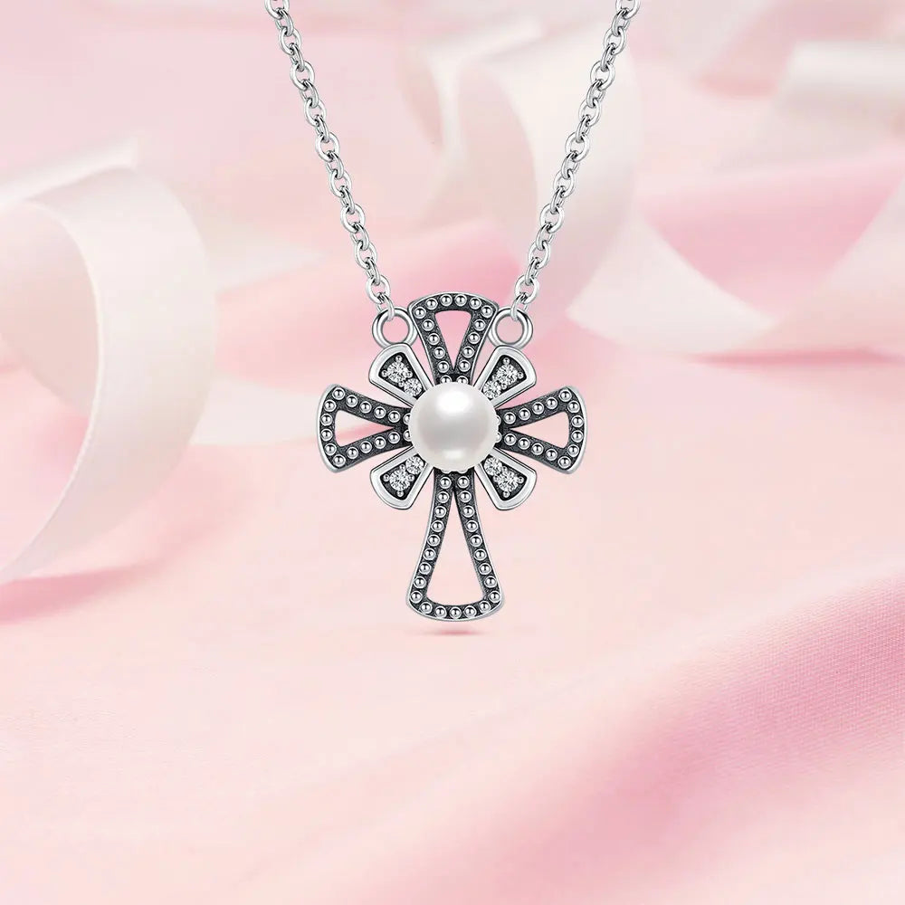 A sterling silver cross pendant necklace with a central white pearl and small clear stones, displayed on a soft pink satin background.