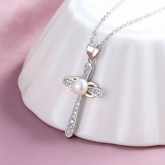 A sterling silver cross white pearl pendant necklace with small clear rhinestones, displayed on a white ceramic stand over a soft pink fabric background.