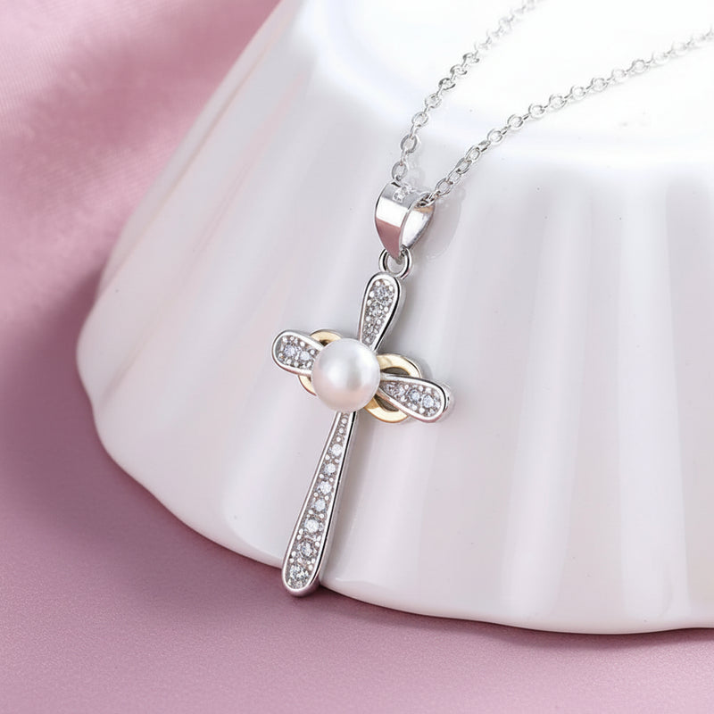 A sterling silver cross white pearl pendant necklace with small clear rhinestones, displayed on a white ceramic stand over a soft pink fabric background.