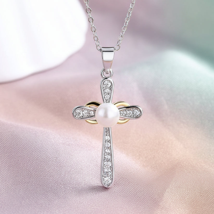 A silver cross pendant necklace with a central white pearl and clear rhinestone accents, displayed on a draped pastel satin fabric background.