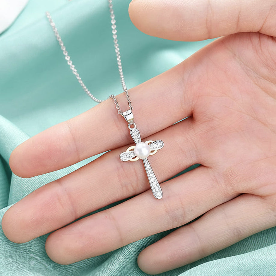 A silver cross pendant with clear stones, displayed on a person's hand over a draped aqua fabric background.