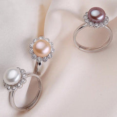 Three sterling silver rings featuring white, pink, and purpler freshwater pearls with sparkling gemstone flower halos displayed on a soft fabric background.