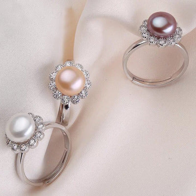 Three sterling silver rings featuring white, pink, and purpler freshwater pearls with sparkling gemstone flower halos displayed on a soft fabric background.