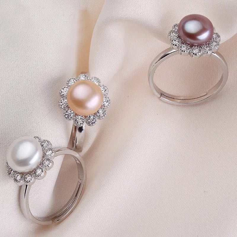 Three sterling silver rings featuring white, pink, and purpler freshwater pearls with sparkling gemstone flower halos displayed on a soft fabric background.