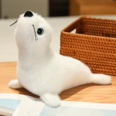 White sea lion plush with black whiskers, a black nose and blue-rimmed eyes beside a wicker basket.